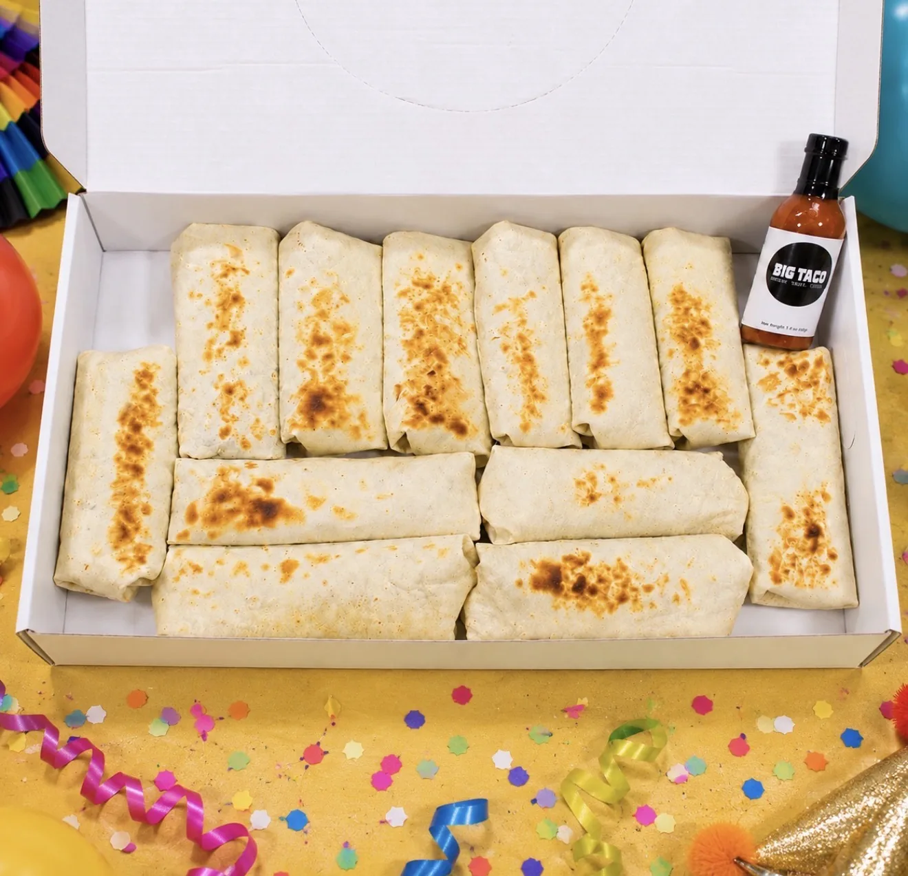 A Big Taco catering box filled with twelve wrapped burritos and a bottle of Big Taco hot sauce, on a party table with confetti and balloons