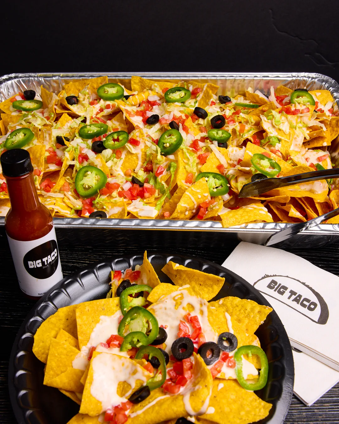 A catering tray of Big Taco loaded nachos with serving tongs, a plated helping in front, a bottle of Big Taco hot sauce and a Big Taco napkin