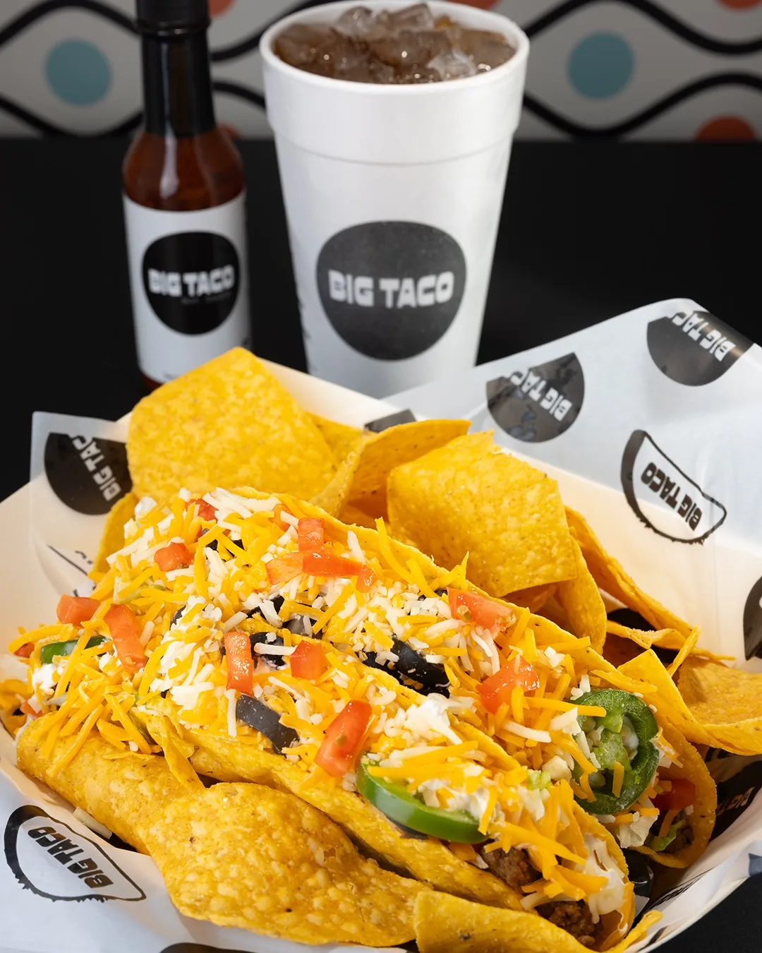Big Taco tacos loaded with cheese, tomato and jalapeno, with tortilla chips, a bottle of Big Taco hot sauce and a drink